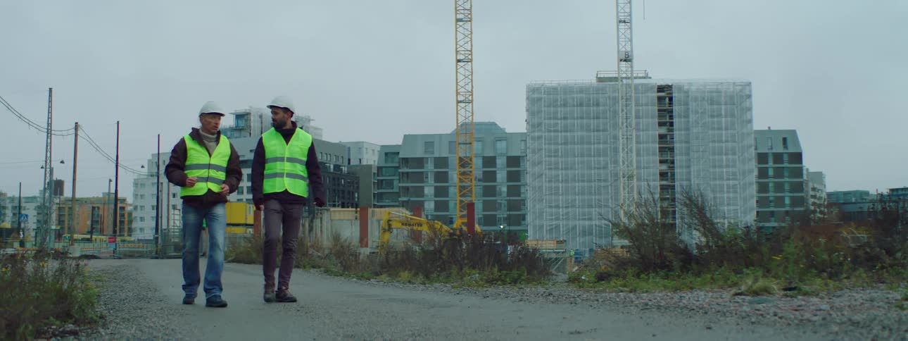 Two construction workers are walking outside in front of buildings and a crane and pointing at something as they talk