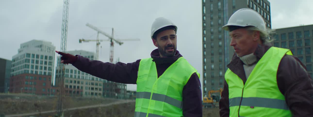 Two male construction workers wearing safety helmets and green vests walking and talking together at a construction site with buildings at the background