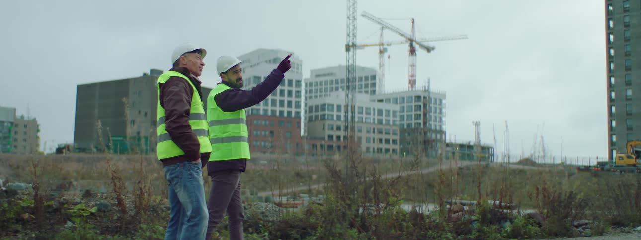 Two male construction workers wearing safety helmets and vests walking and talking together at a construction site with buildings and construction cranes at the background