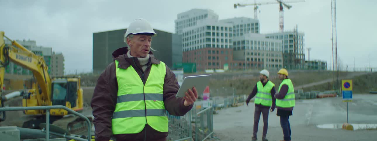 A construction worker standing in a construction site wearing safety helmet and green vest video calling someone on his tablet while two other construction workers are talking at the background