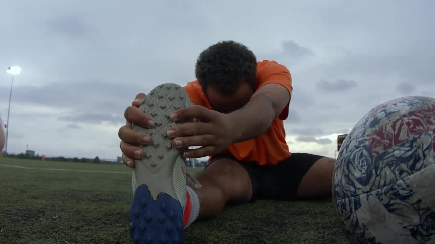 A man sitting on a soccer field stretching his hamstring muscle leaning over his leg holding his cleat