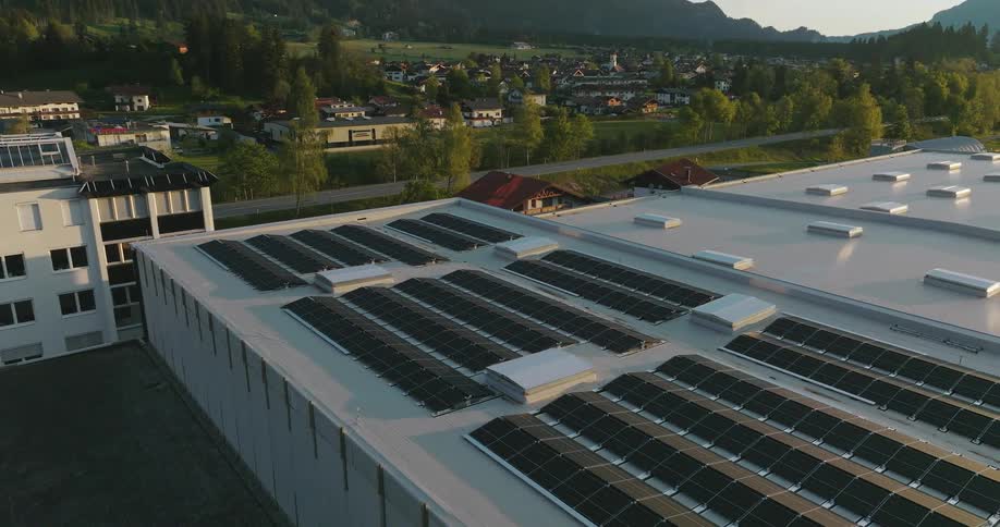 Solar panels are on the roof of a building and neighborhood houses and trees are in the distance