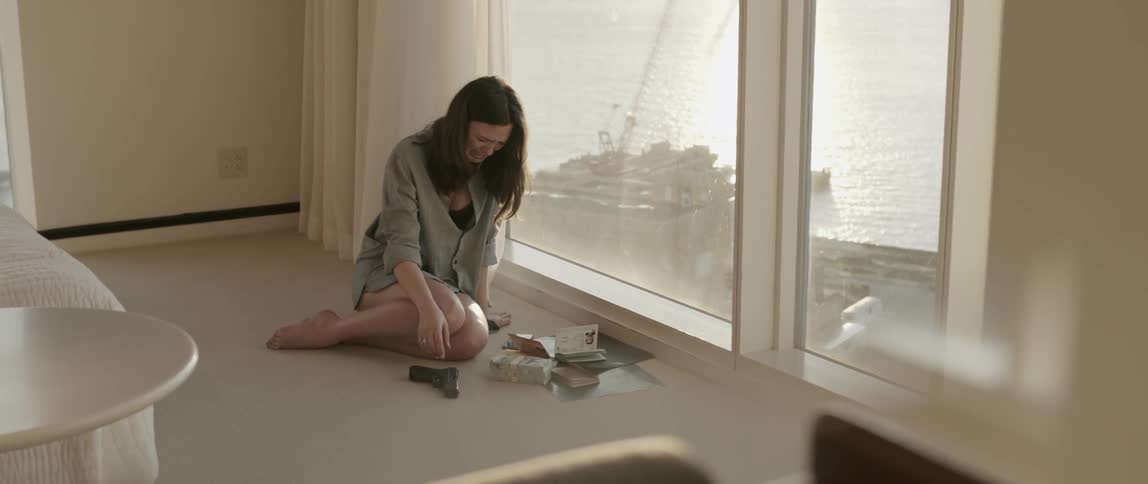 A woman sits on the floor near a window crying and picking up a gun