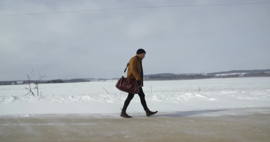 A man is walking down a road with a snow covered field behind him
