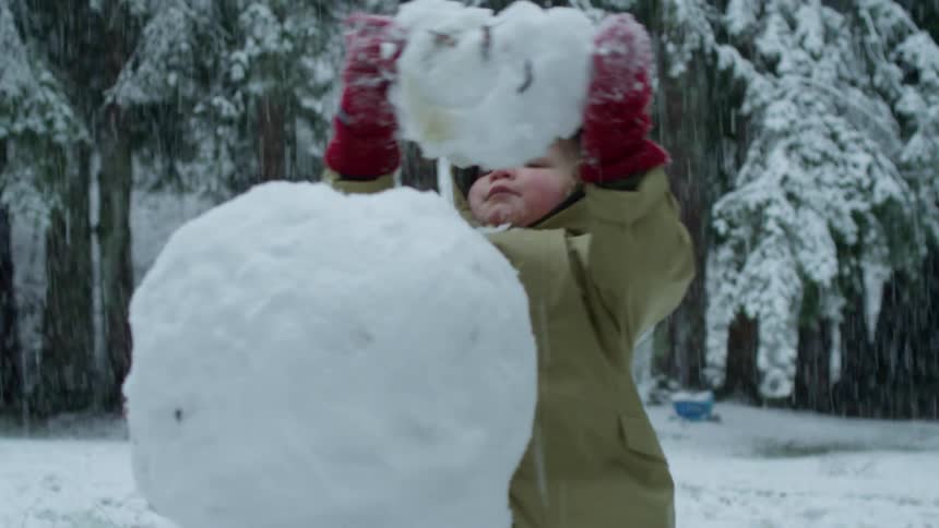 Snow falls as a young child is outside building a snowman
