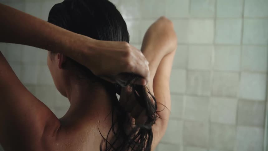 A woman is standing in the shower and moving her hands down her long dark wet hair