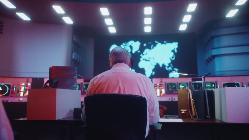 A man sitting working at a table in a control room talking on a headset