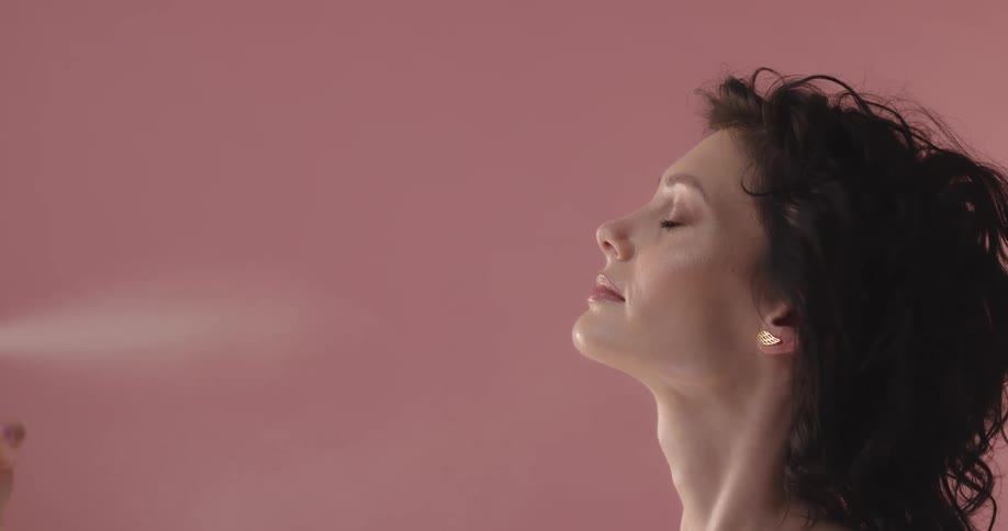 A woman spritzes her face with water in a studio with pink walls