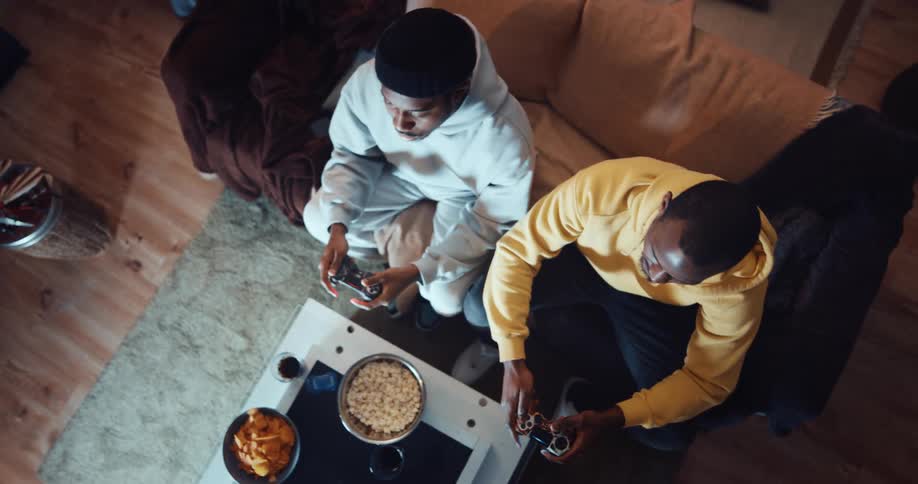 An aerial view of two men sitting at home and talking and laughing as they play a video game