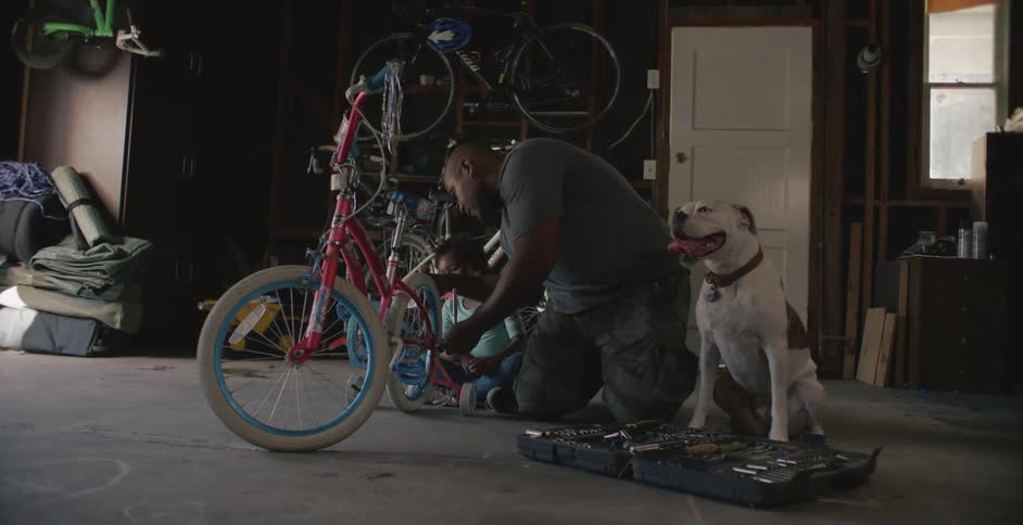 A man tightens the training wheels on the bicycle of a young girl sitting beside him in a garage during the day
