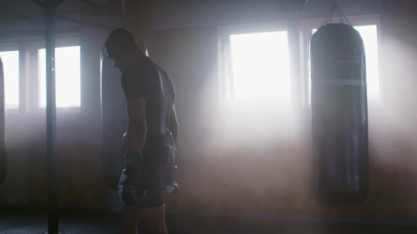 A man wearing boxing gloves and a belt paces in a gym near punching bags