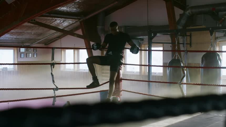Male boxer with boxing gloves trunks and shoes rests in the corner of boxing ring