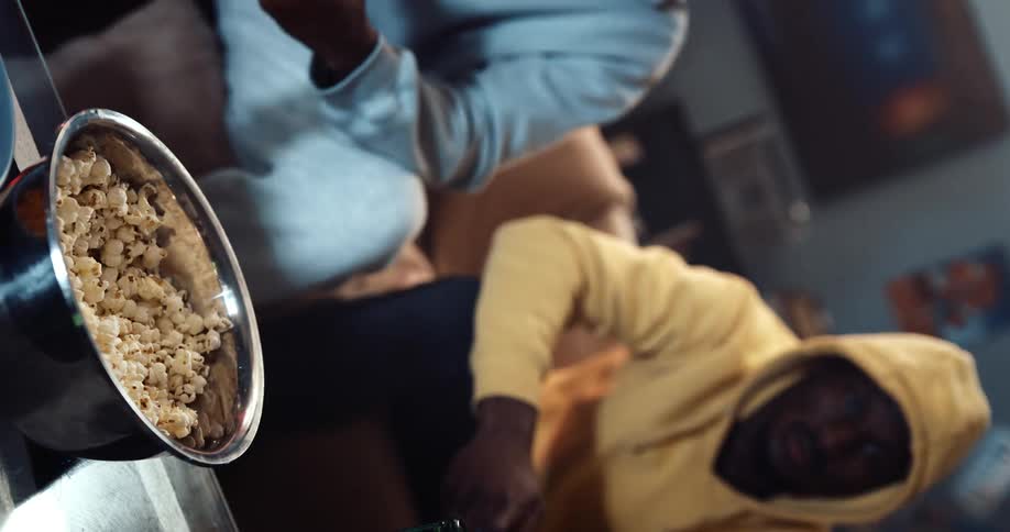 Men with gray and yellow hoodies eating popcorn and drinking from bottle while playing video game at home