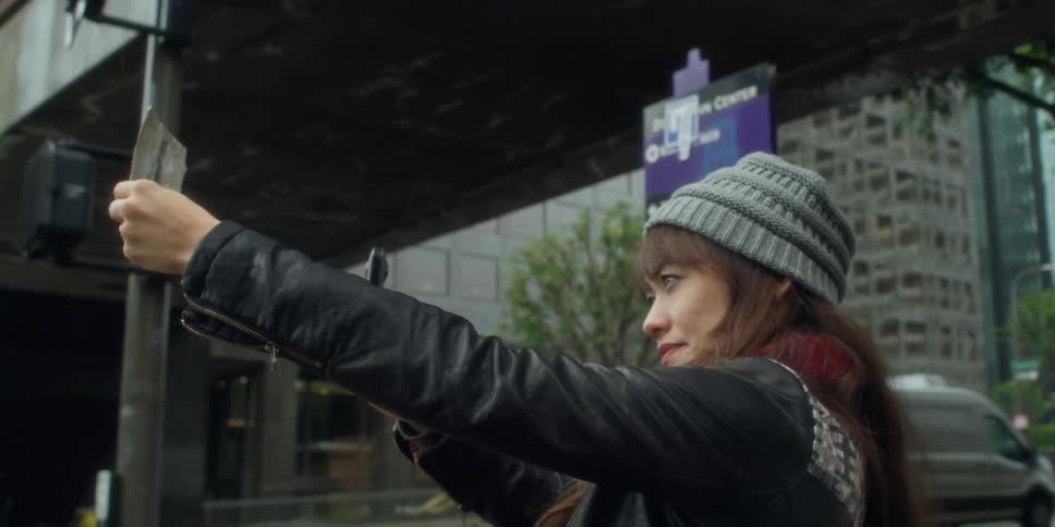 A woman is standing on a busy downtown street holding up a photograph and her phone