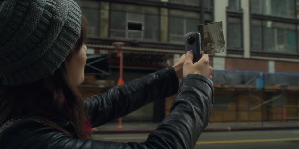 A woman standing on a city sidewalk holds a photograph in front of her and takes a picture of it