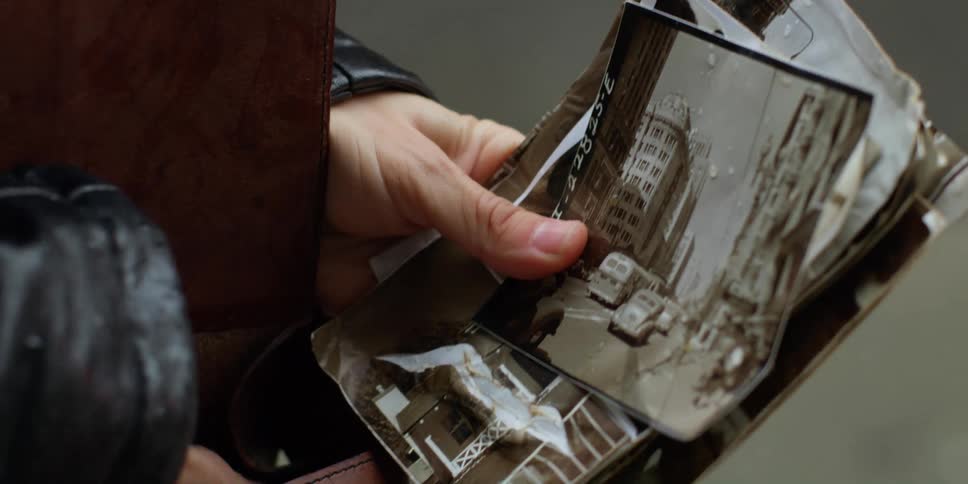 A woman holds a stack of photos with buildings and vehicles and takes a camera out of her pocket