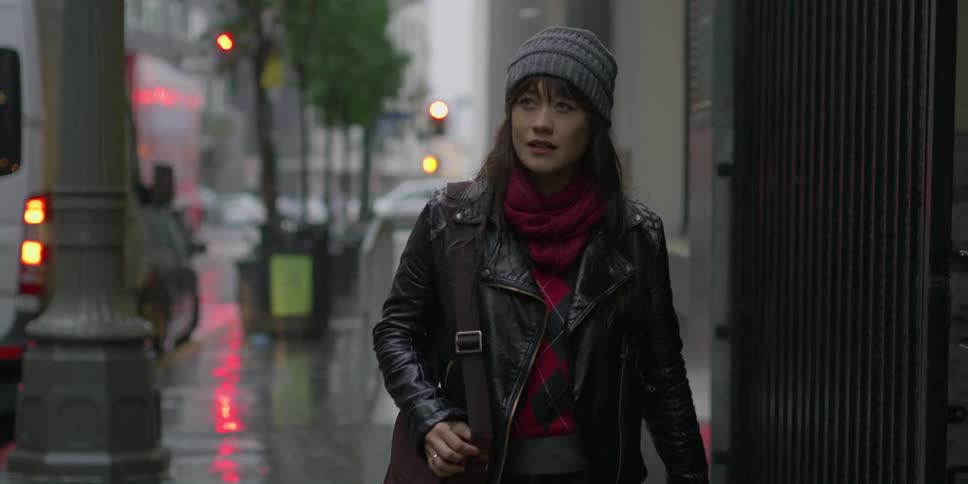 A woman looks up at buildings as she steps out in to the rain and walks down the sidewalk