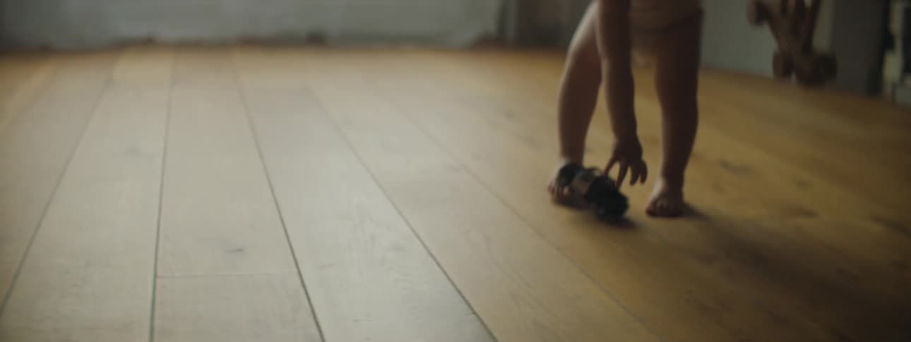 A baby walking in a room picks up a toy and then puts a toy car on the floor and pushes it