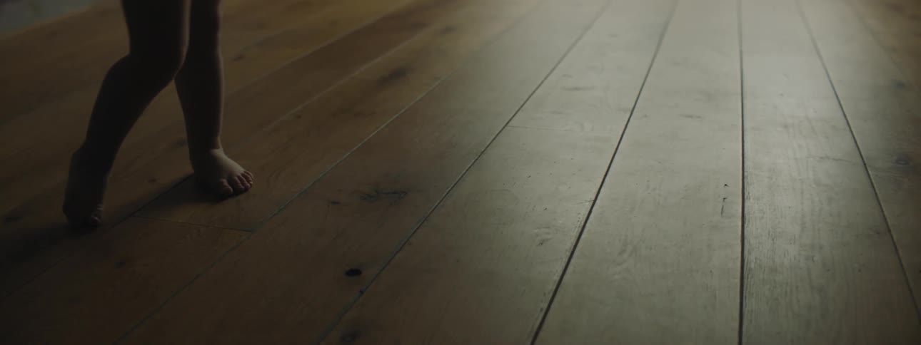 Toddler with bare feet stands in shadow on hardwood floor beside sunlight that is coming in squats down to pick something up then stands up and puts knees together