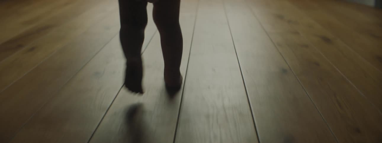 A baby walking on a wooden floor towards a window with curtains