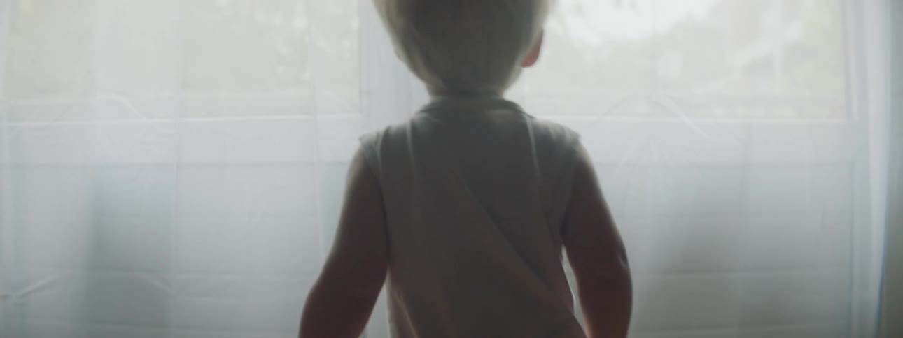 A blonde toddler stands on furniture to look out a window covered with sheer white curtains