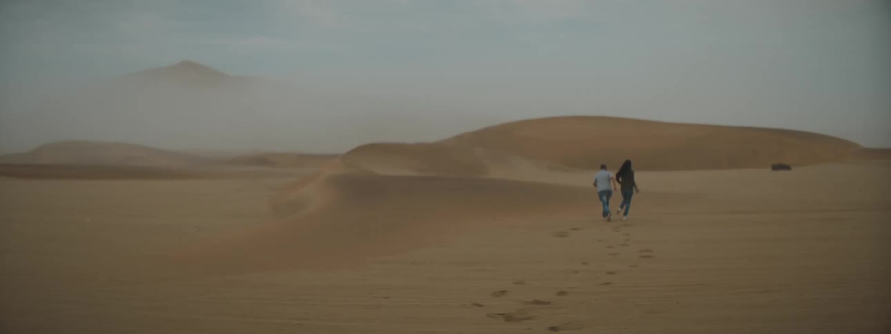 A man and a woman playing tag in a desert with sand dunes the sky and clouds in the background