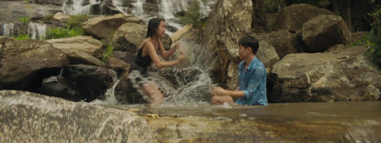 Man and woman play in pool near base of waterfalls flowing down mountain slope