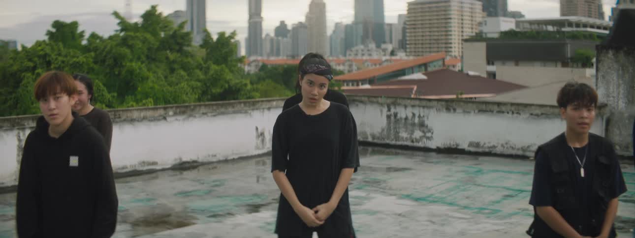 A group of young men and a woman stand on a rooftop near downtown and perform a choreographed dance