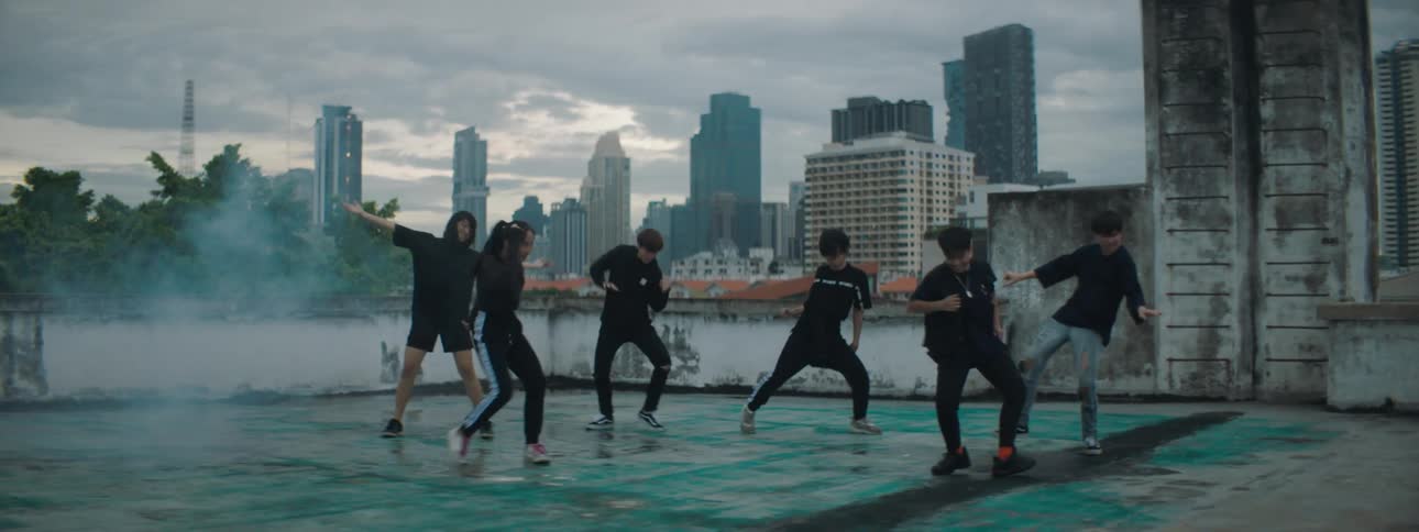 A group of teenagers dance on a rooftop with the city skyline in the distance