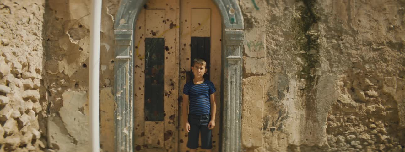 A boy looks into the camera while standing in front of a door in an old stone wall on a sunny day