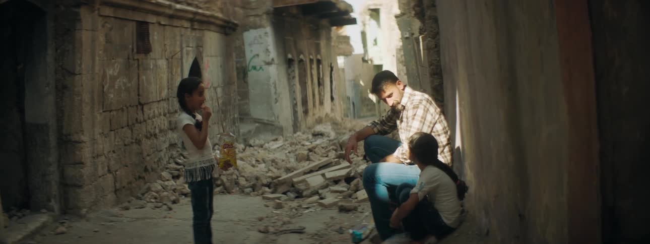 A man sitting in an alleyway between ruined buildings strokes the hair of the girl sitting next to him and talks to her as another girl watches while eating potato chips
