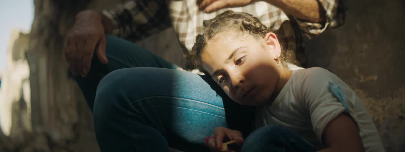 Adult hugs girl then strokes her head while sitting near wall in partly shadowed area