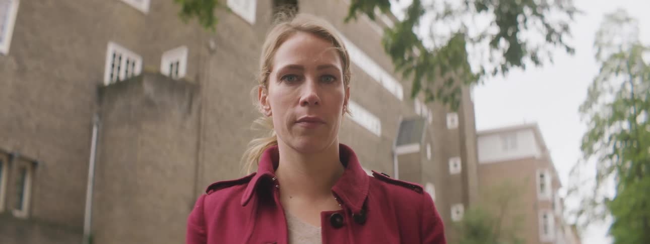 A woman wearing a red jacket stares into the camera while standing outdside of a row of residential buildings during the day