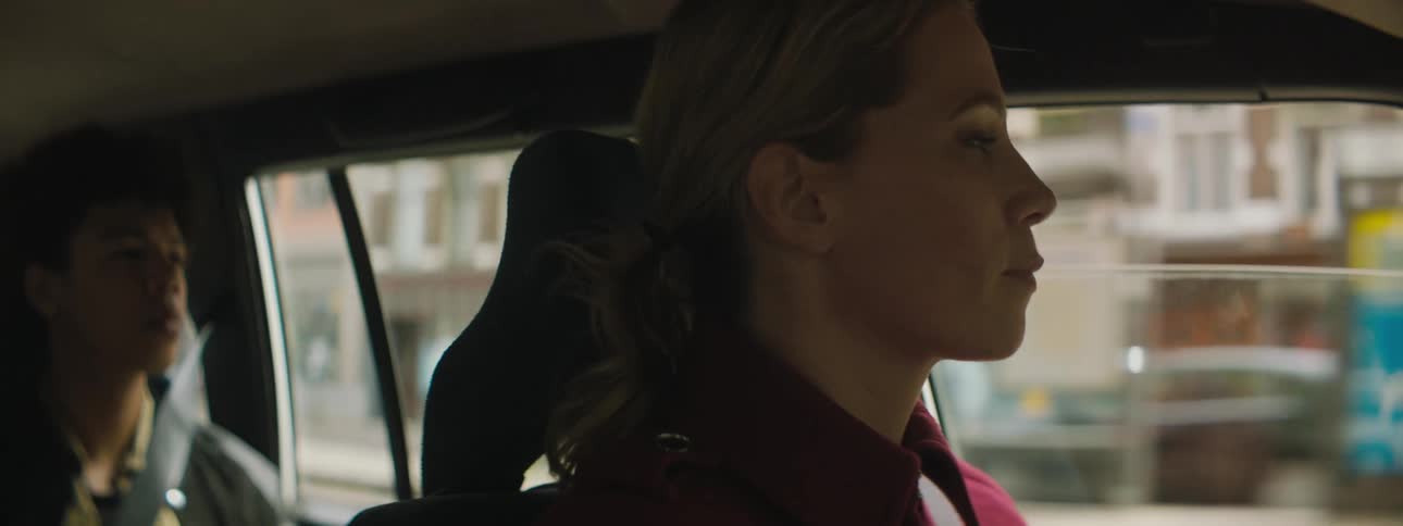 A woman drives a car through a city with buildings and people walking by while a passenger sits in the backseat