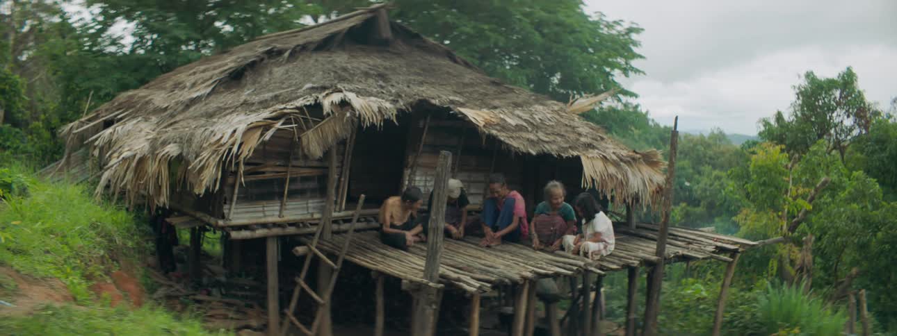 Grandmother talks to girl sitting with rest of family on platform near thatched hut on slope near wilderness