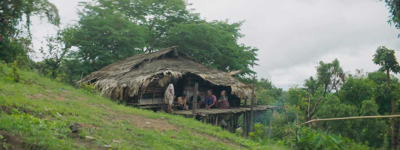 A girl climbs up a ladder onto the porch of a shack on a hill to join her family that is already sitting there