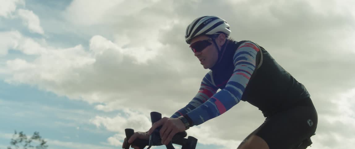 A man wearing a bicycle helmet and sunglasses is riding a bike under a cloudy sky