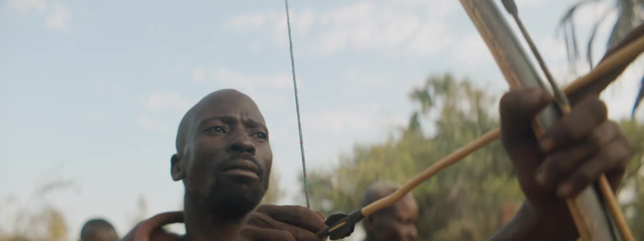 An african american man holding a bow and arrow stands alert looking ahead in a tropical area