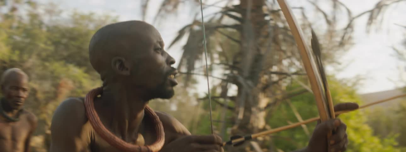 Men in a tribe walk in a forest as one man aims his bow and arrow