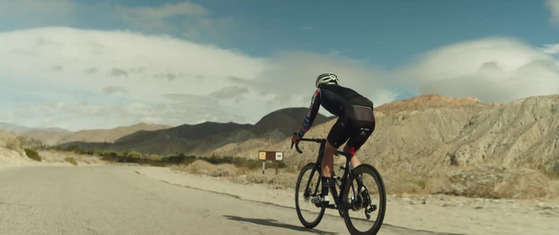 A man wearing a bicycle helmet passes a sign on the roadside as he rides his bike on a dirt road near mountains