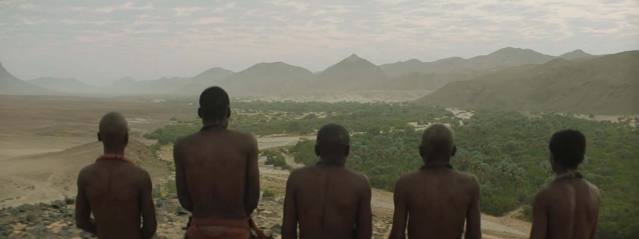 A group of black men in tribal wear stand at high altitude and look out over a foggy countryside that changes goes mountain to forest to desert