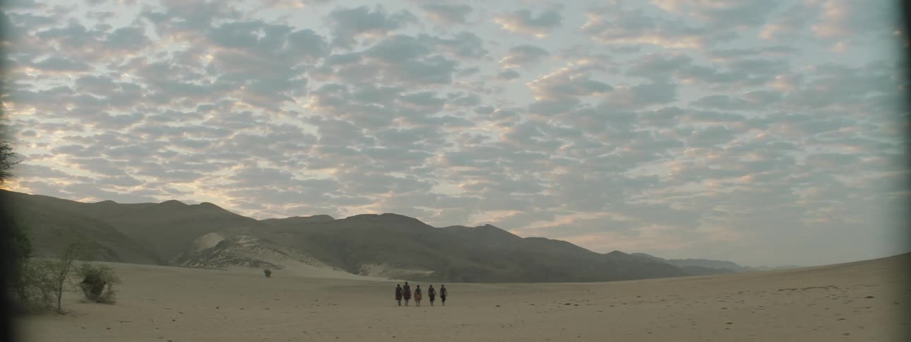 Group of men in traditional attire walk across desert bordered by mist covered mountain under slightly pink cloudy sky