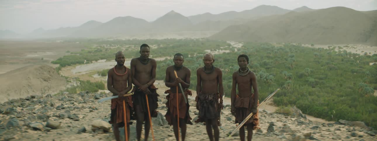A group of black tribal men look into the camera while standing on a cliff overlooking a vast countryside
