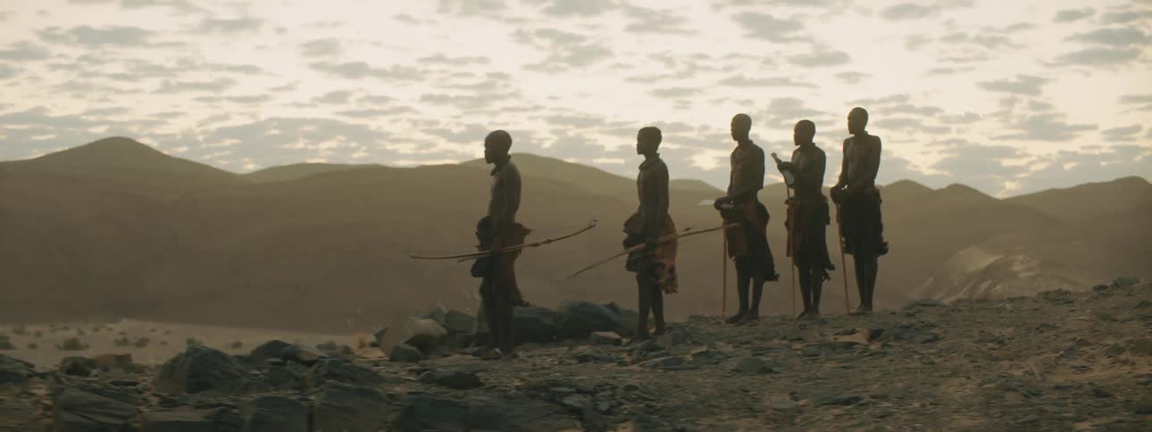 Five shirtless tribesmen stand in a desert near a mountain range at dusk