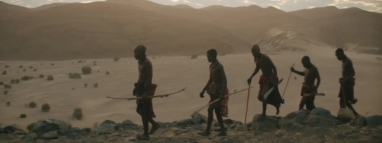 Men walk along rocky ground carrying bows across from misty mountain and sandy valley