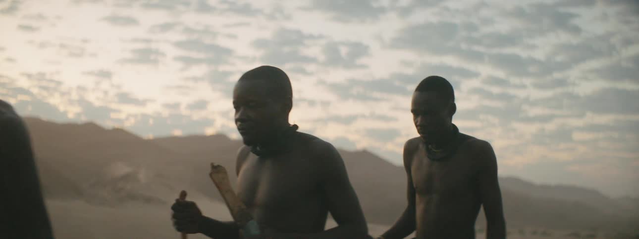 Four people walk past in shadow with sunrise or sunset behind them creating a orange and blue sky full of fluffy dark clouds with hills in the distance