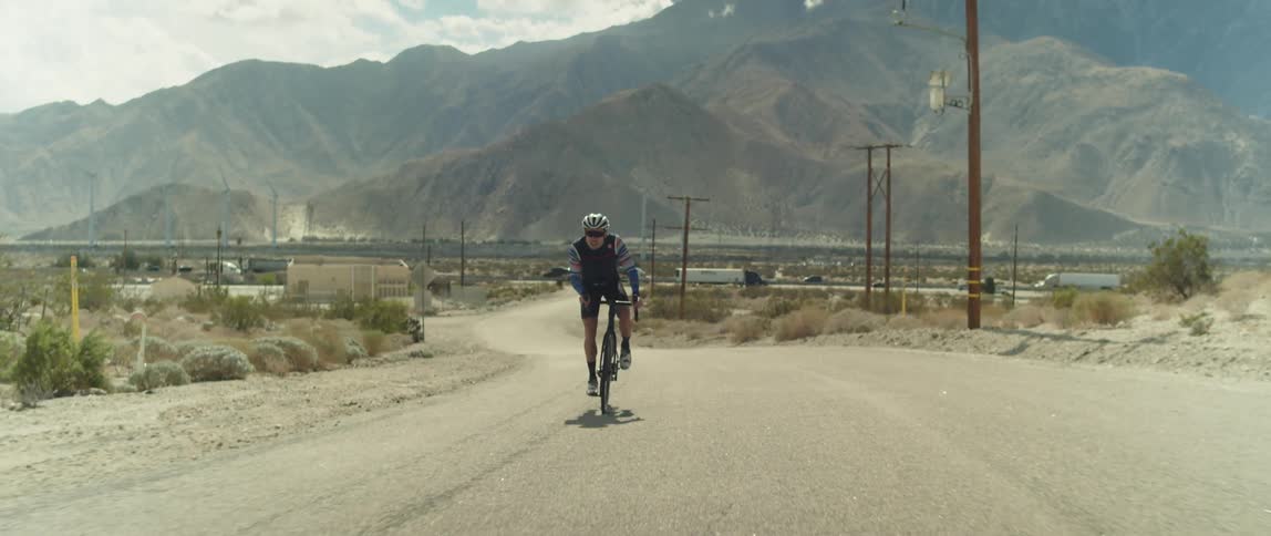 A man cycles on a gravel road with shipping truck and vehicles driving in front of a mountain range behind him