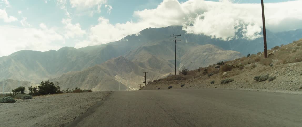 A dirt road is lined with utility poles and mountains are in the distance under a cloudy sky