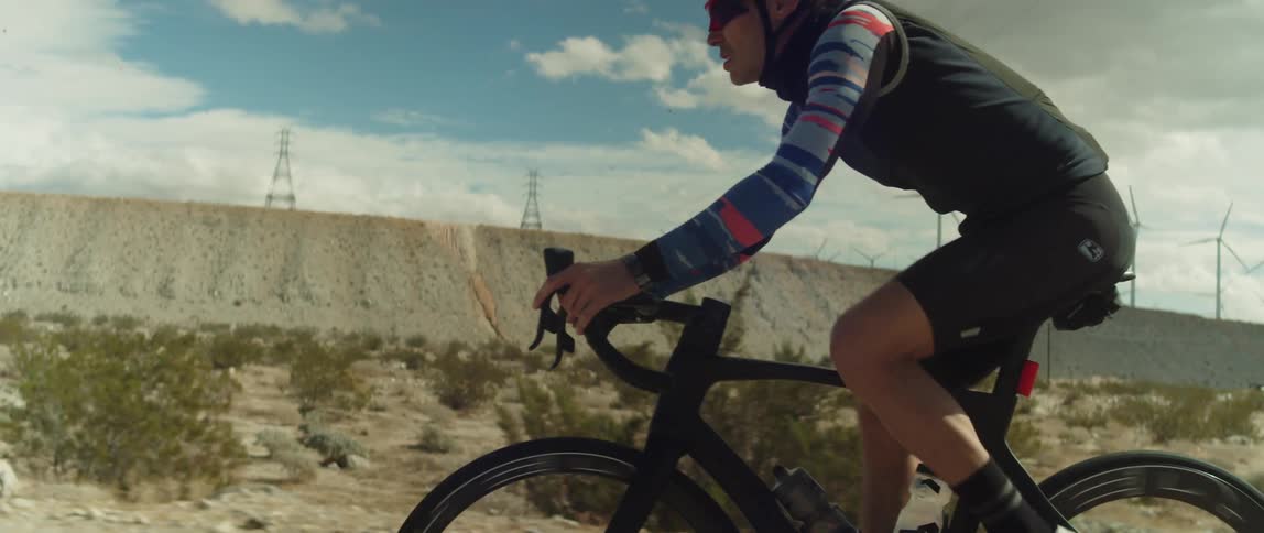 A man wearing a helmet and sunglasses is riding a bike past utility towers and wind turbines on a wind farm