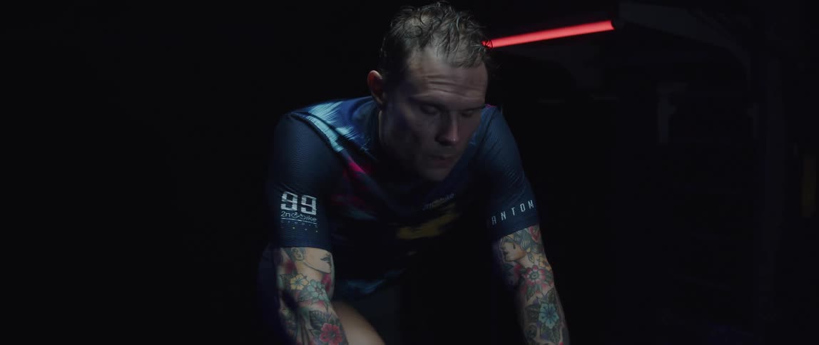 Focused cyclist man wearing a cycling jersey exercising on a stationary bike on a dark room